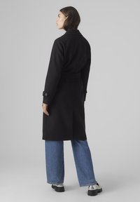 Cappotto di lana nero con cintura, colletto grande e polsini con bottoni, abbinato a jeans in denim a gamba larga e scarpe con platform metallizzate.