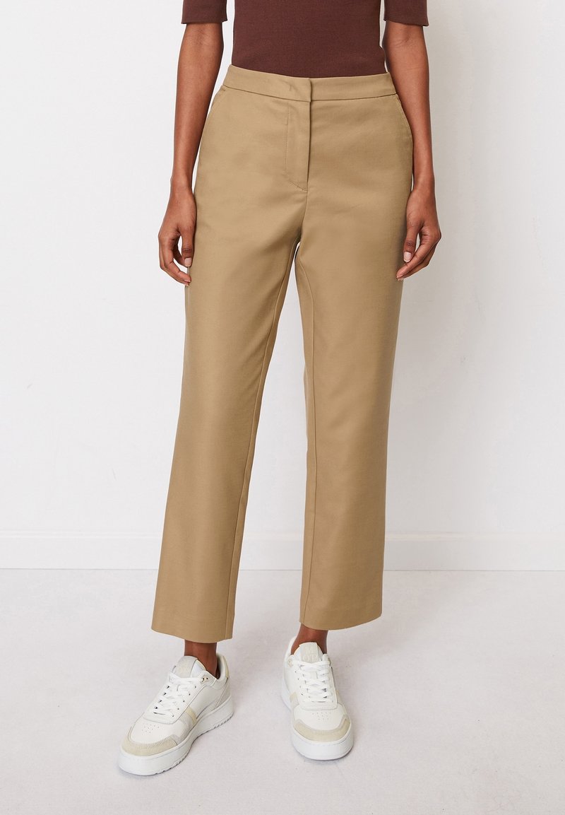 Personne portant un pantalon beige à jambes droites, des baskets blanches et un haut marron à manches longues, se tenant contre un mur blanc.