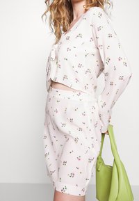 Blusa corta con estampado floral en rosa suave, combinada con pantalones cortos a juego. La modelo sostiene un pequeño bolso verde texturizado.