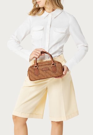 Mujer con camisa blanca de manga larga y pantalones cortos beige sosteniendo un pequeño bolso marrón texturizado con dos asas.