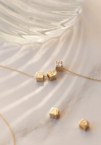 Gouden kettinghalsketting met kubieke schakels met gegraveerde letters en een heldere edelsteenaccent. Glad oppervlak met subtiele reflecterende eigenschappen.