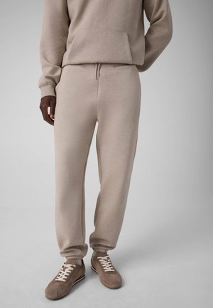 Persona che indossa pantaloni da tuta beige e felpa con cappuccio abbinata con sneakers casual marroni su uno sfondo chiaro uniforme, in piedi con una mano dietro la schiena.