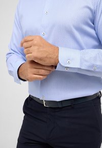 Lichtblauw gemusterd overhemd met een knoopsluiting bij de kraag en manchetten; gecombineerd met donkere broek en een zwarte riem met een zilveren gesp.