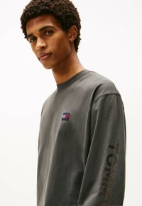 Camisa gris de manga larga con un pequeño logo de color en el pecho y el texto "TOMMY" en grande bajando por una manga. Tela de algodón suave, corte relajado.
