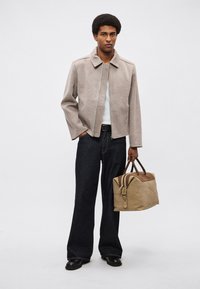 Jasnobrązowa, teksturowana kurtka z kołnierzem, biała koszula, ciemne dżinsy oraz beżowa torba podróżna z ciemnymi akcentami. Czarny obuwie dopełnia strój.