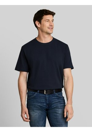 Man draagt een effen donkerblauw T-shirt met korte mouwen, ingestopt in blauwe spijkerbroek met een zwarte riem, kijkt opzij en glimlacht iets.