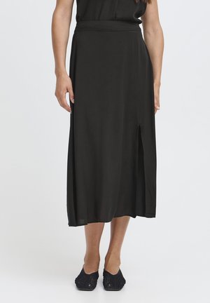 Naine musta poolpika seeliku ja külje lõhikuga, kandes musti libisemisjalanõusid, seisab tavalise heleda tausta ees.