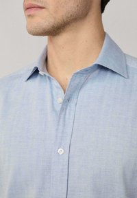 Camicetta azzurra chiara, realizzata in tessuto intrecciato con un sottile motivo a spina di pesce. Presenta un colletto classico e bottoni bianchi.