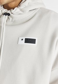 Felpa grigia chiara con tessuto testurizzato, dotata di chiusura con lacci e un distintivo patch logo in bianco e nero sul petto.