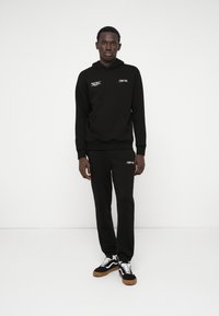 Conjunto de hoodie e calças de moletom pretas, feito de tecido macio, com um bolso frontal, punhos elásticos e uma marcação branca no peito e na coxa.