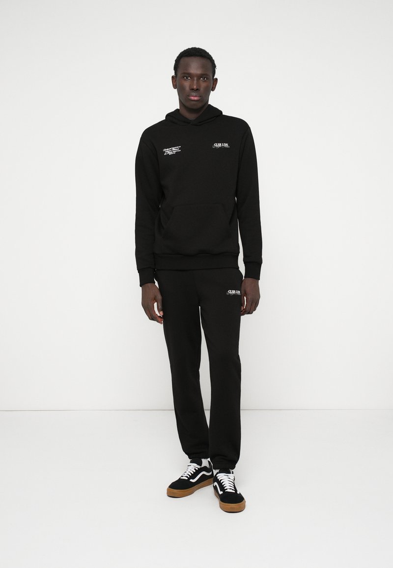 Conjunto de hoodie e calças de moletom pretas, feito de tecido macio, com um bolso frontal, punhos elásticos e uma marcação branca no peito e na coxa.