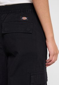 Gros plan d'une personne portant un pantalon cargo Dickies noir avec une taille élastique et des poches à rabat, main reposant sur le côté.