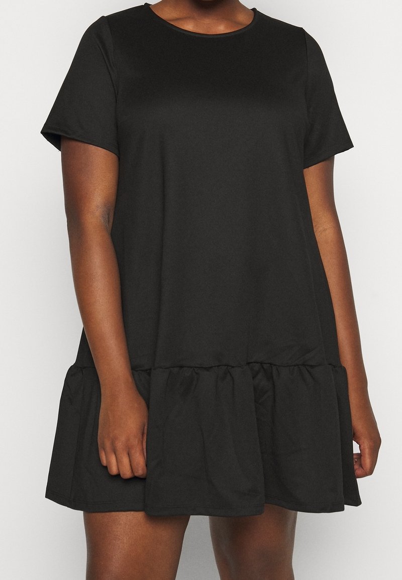 Femme portant une robe noire à manches courtes, coupe ample et ourlet à volants, debout devant un fond uni.
