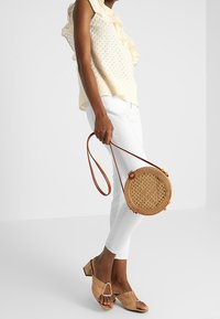 Kvinna i krämfärgad blus med volanger, vita smala byxor, bruna sandaler med öppen tå och klack, som håller en rund, vävd axelremsväska med läderrem.