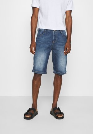 Mann trägt mittellange blaue Jeansshorts, weißes T-Shirt und schwarze Sandalen mit Schnalle, steht vor einem weißen Hintergrund.