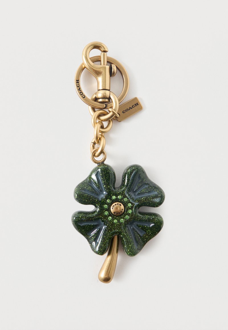 Porte-clés en métal doré avec un charm trèfle à quatre feuilles scintillant vert foncé et de petits strass verts, marqué avec des étiquettes « Coach ».