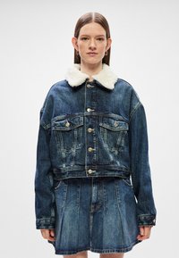 Τζάκετ από τζιν με πουκάμισο από γούνα, φθαρμένο μπλε ύφασμα, χρυσά κουμπιά και δύο τσέπες στο στήθος. Συνδυασμένο με φούστα από τζιν.