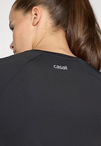 Svart träningsskjorta med slät textur, försedd med rundad halsringning och "casall"-logotypen i vitt på övre delen av ryggen.