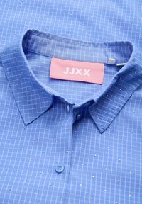 Camisa a cuadros azul claro con detalles metálicos, cuello italiano y cierre de botones. Etiqueta de la marca "JJXX" en rosa en el cuello.