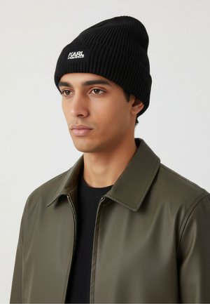Schwarze gerippte Strickmütze mit weißem "KARL LAGERFELD" Logo auf der Vorderseite; getragen mit einem schwarzen Hemd und einer olivgrünen glänzenden Jacke.