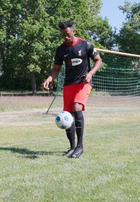 Tricou de fotbal cu mânecă scurtă, negru, cu accente roșii și logo, șorturi roșii, șosete negre; jucător driblând o minge de fotbal cu model pe iarbă.