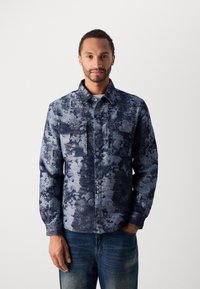Blue patterned jacket with two chest pockets, button front, long sleeves, and a textured fabric. Worn over a light shirt and jeans.
