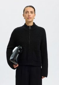 Juodas pūkuotas kardiganas su aukštu apykakliu, užtrauktuku ir priekinė kišene. Derinamas su juoda struktūruota rankine. Modelis vilki tamsias kelnes.