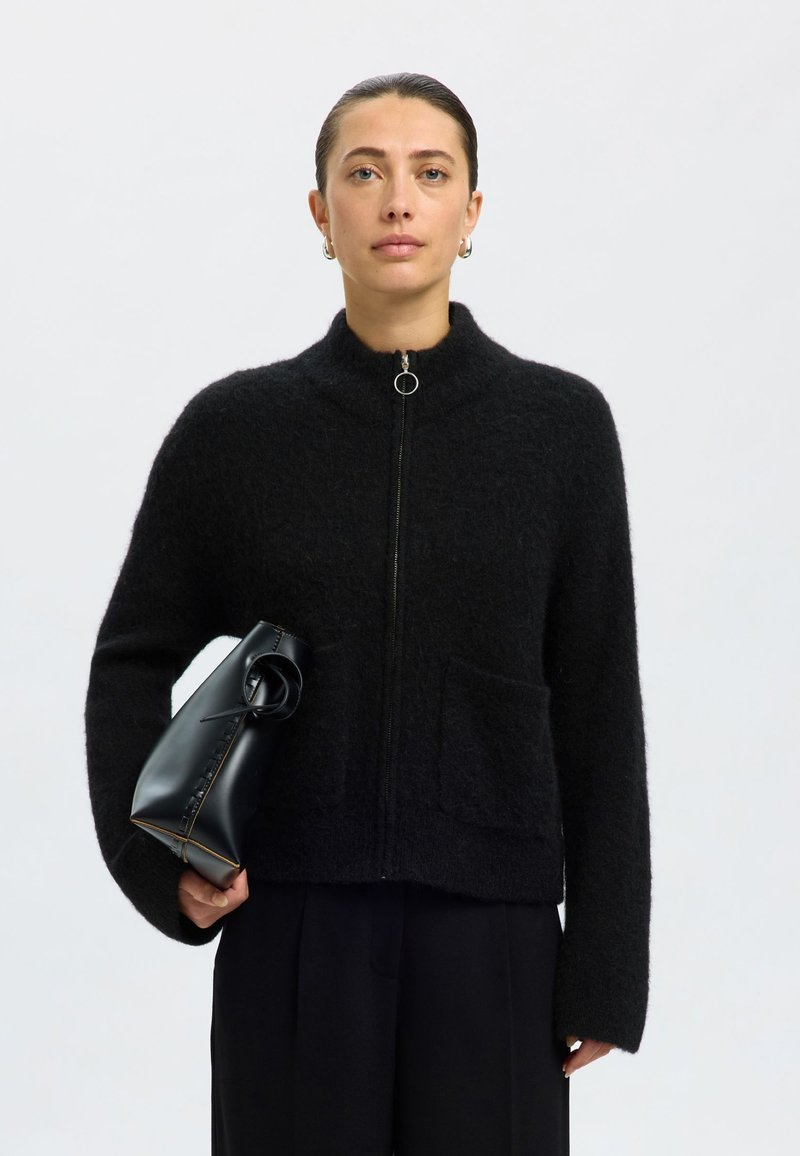 Juodas pūkuotas kardiganas su aukštu apykakliu, užtrauktuku ir priekinė kišene. Derinamas su juoda struktūruota rankine. Modelis vilki tamsias kelnes.