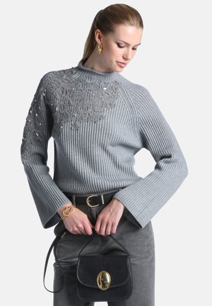 Femme portant un pull côtelé gris avec des détails en dentelle et paillettes, un pantalon gris, des bijoux en or, et tenant un sac à main noir avec un fermoir doré.