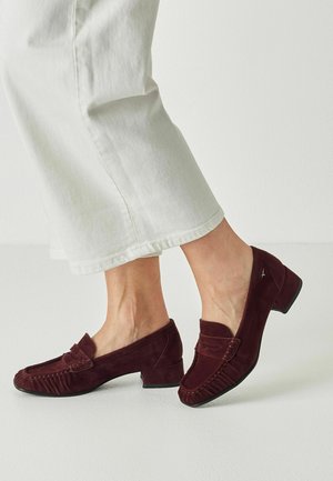 Person iført hvide korte bukser og mørkerøde ruskind lave hælede loafers, stående på en lys gulv mod en hvid væg.