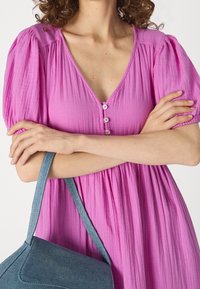 Rosa texturerad klänning med V-ringning och korta puffärmar, utrustad med en rad knappar, i kombination med en blå denimväska.