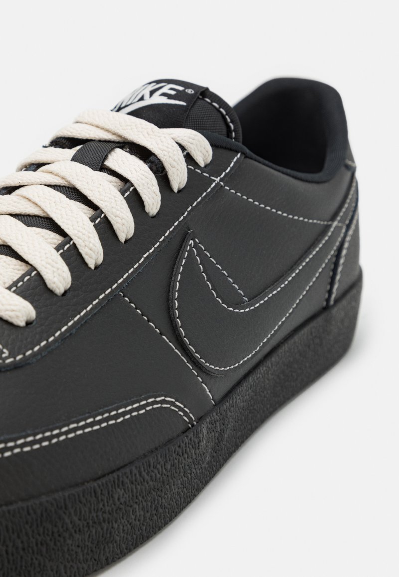 Zapatillas de cuero negro con cordones crema, costuras en contraste y suela de goma negra texturizada. Presenta un logotipo prominente en la lengüeta.