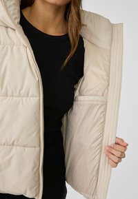 Beige pufferväst med dragkedja och strukturerad detalj, med en mjuk innerfoder och en innerficka på ena sidan.