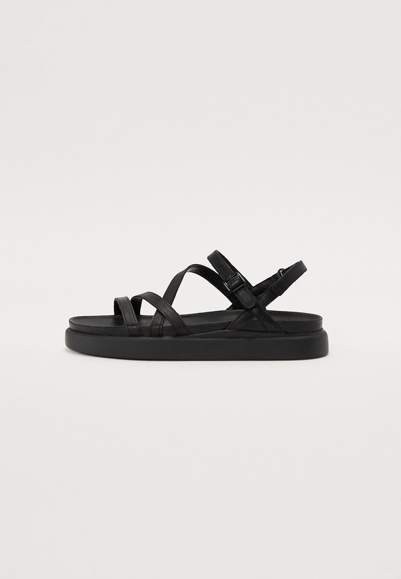 Sandales noires avec une semelle plate et texturée, dotées de brides croisées et d'une sangle à boucle ajustable sur le côté.