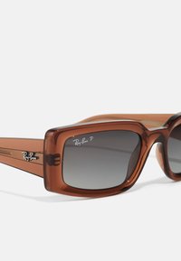 Barna, túlméretezett napszemüveg vastag kerettel, sötét árnyalatú, fokozatosan sötétedő lencsékkel és ezüst Ray-Ban logóval az elején és a szárakon.