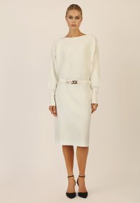 Weißes, geripptes Strickkleid mit weitem Ausschnitt, langen Ärmeln und einem taillierten Gürtel, der mit einem glänzenden silbernen Kettendetail verziert ist. Schwarze Absatzschuhe.