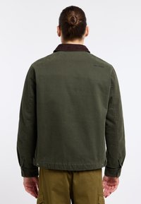 Green canvas jacket with a brown corduroy collar, visible stitching, and buttoned cuffs. Back view shows a simple design and subtle branding.
