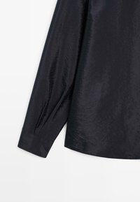 Chemise noire en satin à manches longues avec poignets boutonnés et un léger éclat sur tissu lisse sur fond blanc.