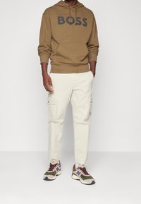 Felpa con cappuccio marrone con la scritta "BOSS" in nero, abbinata a pantaloni cargo beige chiaro con tasche laterali e sneakers multicolori.