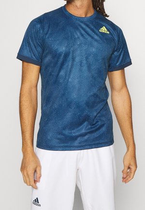 Mężczyzna w niebieskiej sportowej koszulce Adidas z subtelnym wzorem i żółtym logo, ubrany w białe spodenki Adidas na jednolitym tle.