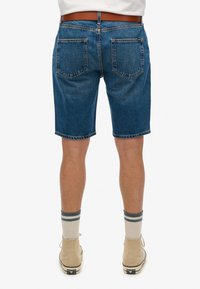 Pantalones cortos de denim en azul, con corte recto, cinco bolsillos y dobladillos deshilachados. Llevados con zapatillas deportivas altas color crema y calcetines a rayas.