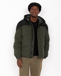 Casaco tipo puffer em verde escuro e preto, com capuz, fecho de zipper e design acolchoado. Usado sobre uma camisa preta com calças castanhas.