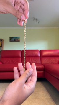 Złoty łańcuszek trzymany w dłoni, z połączonymi segmentami, błyszczącą powierzchnią i małym zawieszkiem na jednym końcu. W tle znajduje się czerwona kanapa.