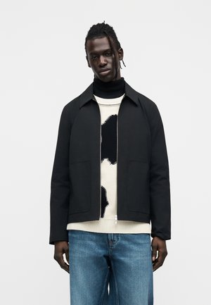 Μαύρο ελαφρύ μπουφάν με κλασικό γιακά, δύο μπροστινές τσέπες, φορεμένο πάνω από ένα υφασματόμενο εκρού πουλόβερ με μαύρο μοτίβο, συνδυασμένο με μπλε τζιν.