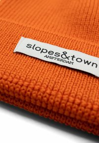 Berretto lavorato a maglia arancione con texture a costine e un'etichetta bianca con la scritta "slopes&town AMSTERDAM" in lettere nere.