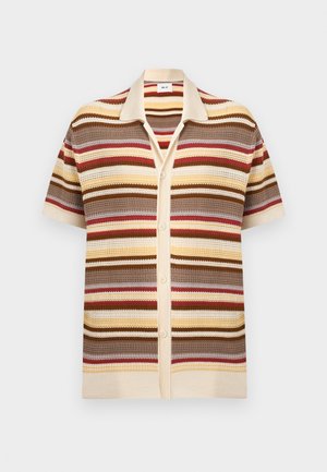 Hombre de pie con las manos en los bolsillos, vistiendo una camisa de botones de manga corta a rayas, pantalones cortos oscuros y sandalias marrones sobre fondo blanco.
