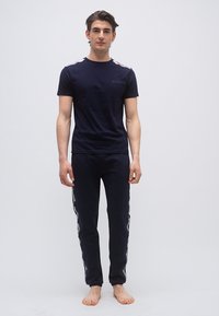 Jeune homme debout pieds nus portant un T-shirt bleu marine et un pantalon assorti avec un ruban à logo blanc sur les épaules et le long des côtés du pantalon.