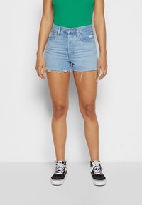 Person som bär ljusblå fransiga denimshorts, grön ärmlös topp och svarta högsko-sneakers med vita snören, står mot vit bakgrund.