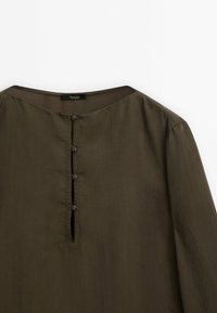 Blouse à manches longues vert olive avec un col rond, dotée d'une patte de boutonnage et d'une texture douce et lisse. Légère brillance et design minimaliste.