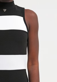 Vestido negro sin mangas con rayas horizontales blancas, textura acanalada, cuello alto y un acento metálico triangular en el cuello.